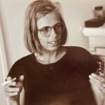 Hilde Wackerhagen sitting in a wicker chair, holding a cigarette in one hand and gesturing with the other; she wears glasses and a dark T-shirt.