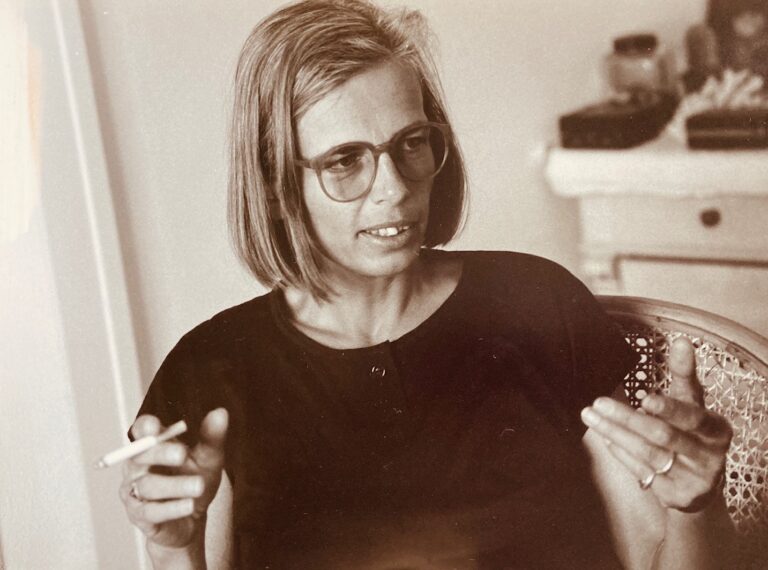 Hilde Wackerhagen sitting in a wicker chair, holding a cigarette in one hand and gesturing with the other; she wears glasses and a dark T-shirt.